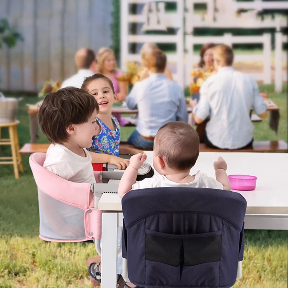 Two toddlers are seated in sturdy, Portable Baby Dining Chairs by Gigi & Jade Kids Co. at an outdoor table, facing each other. One toddler is in a pink chair, and the other is in a navy-blue chair. In the background, several adults are seated around another table, blurred and engrossed in conversation.