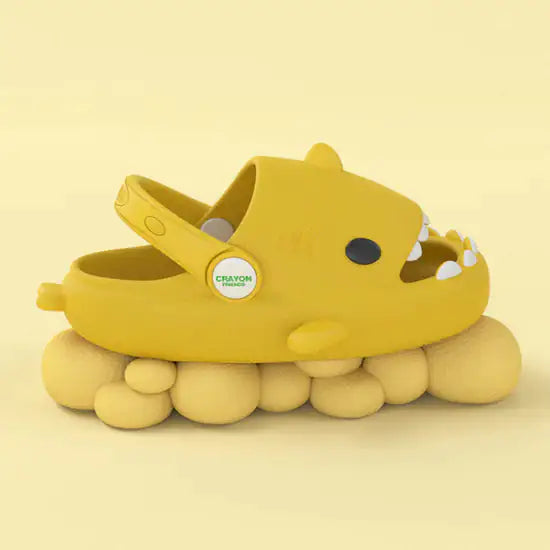 A playful shark-themed kids slipper called "Comfy Sharks with Strap" from Gigi & Jade Kids Co., features a vibrant yellow color with eyes, nostrils, and teeth, mimicking an animal design. The soft yellow slipper rests on a sole that resembles rounded, pebble-like formations.