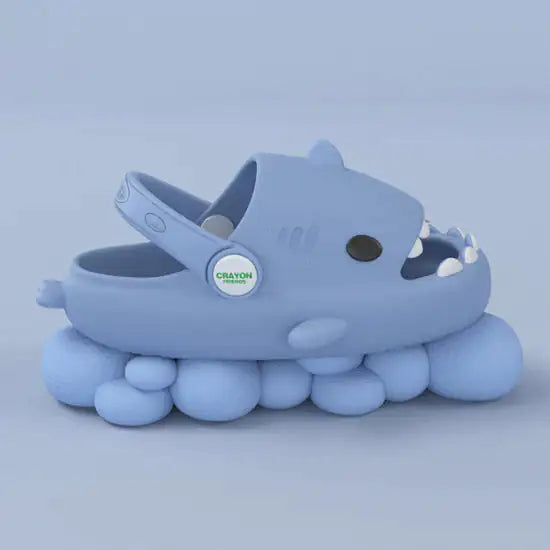 The "Comfy Sharks with Strap" by Gigi & Jade Kids Co. is a blue children's shoe designed to resemble a shark, complete with a rounded body, eyes, and teeth. These charming kids slippers provide cozy comfort thanks to their unique, cloud-like sole made of multiple soft, rounded shapes. A button on the side proudly displays the word "CRAYON.