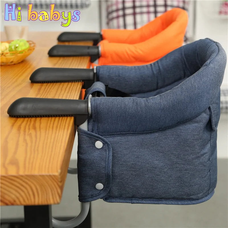 The Portable Baby Dining Chair by Gigi & Jade Kids Co. is designed for babies and securely attaches to the edge of a wooden table. One chair comes in dark blue and another in orange. Featuring cushioned seats and black plastic arms for attachment, these chairs ensure safe seating while "Hi babys" is printed in colorful letters in the top left corner.